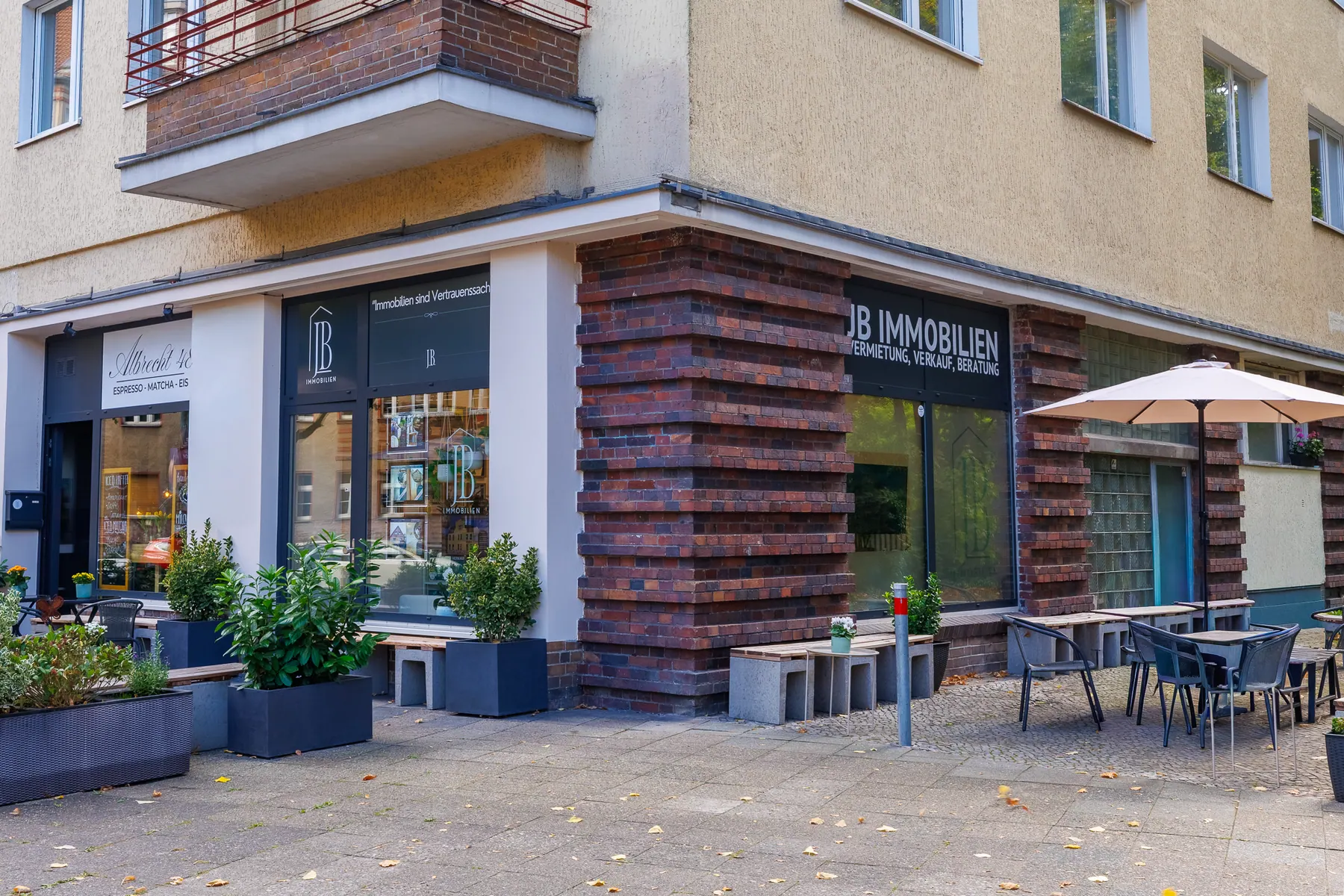 Büro von JB Immobilien an der Straßenecke Albrechtstraße 48, Berlin-Steglitz: Schaufenster mit Logo, Sitzplätze und Sonnenschirm davor